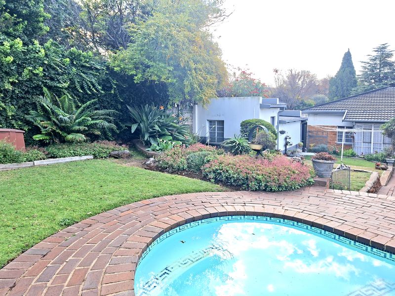 A well-maintained backyard featuring a swimming pool with brick paving, surrounded by established garden beds and mature greenery.
