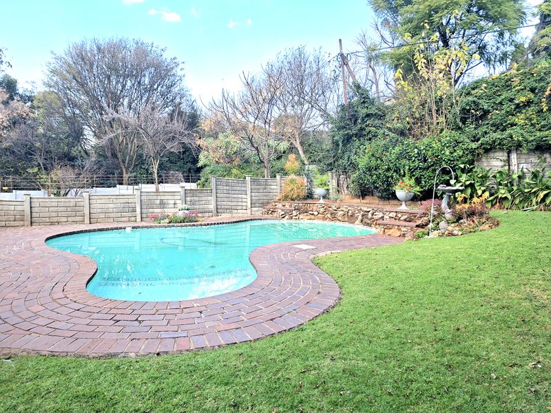 A well-maintained backyard featuring a kidney-shaped swimming pool with a brick surround, stone retaining wall, and established garden landscaping.