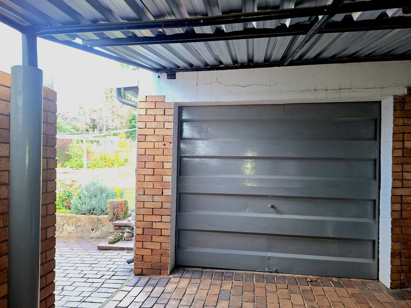 A covered carport area featuring a brick structure, paved flooring, and a grey sectional garage door leading to the rear garden.