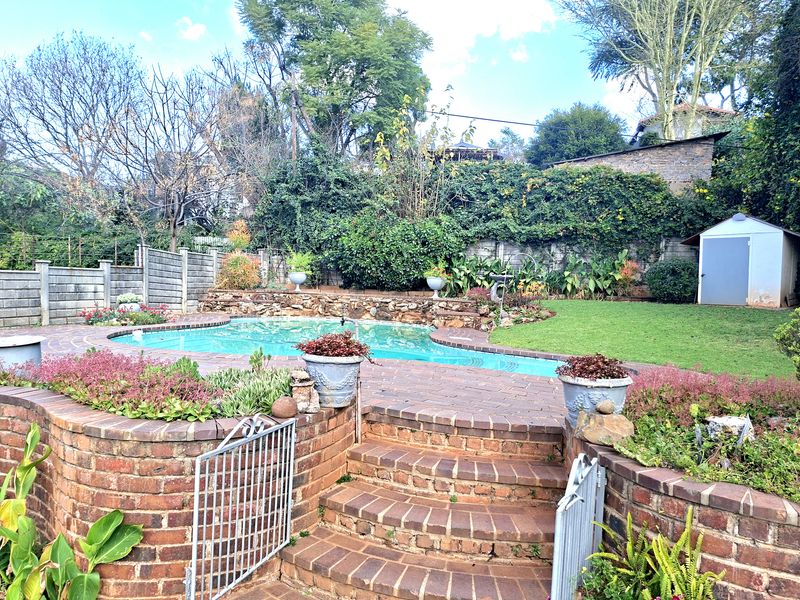 A landscaped backyard featuring a swimming pool, brick-paved patio area with retaining walls, and a garden shed set within a mature garden.