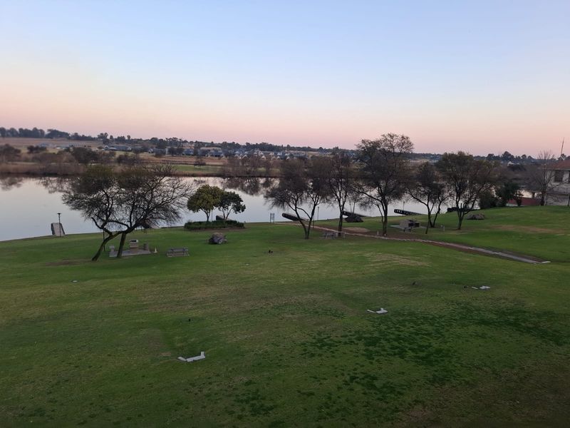 A scenic elevated view overlooking a well-maintained green lawn, mature trees, and a tranquil lake at sunset.