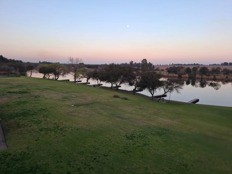 A scenic elevated view overlooking a tranquil river, landscaped lawns, and mature trees at sunset.