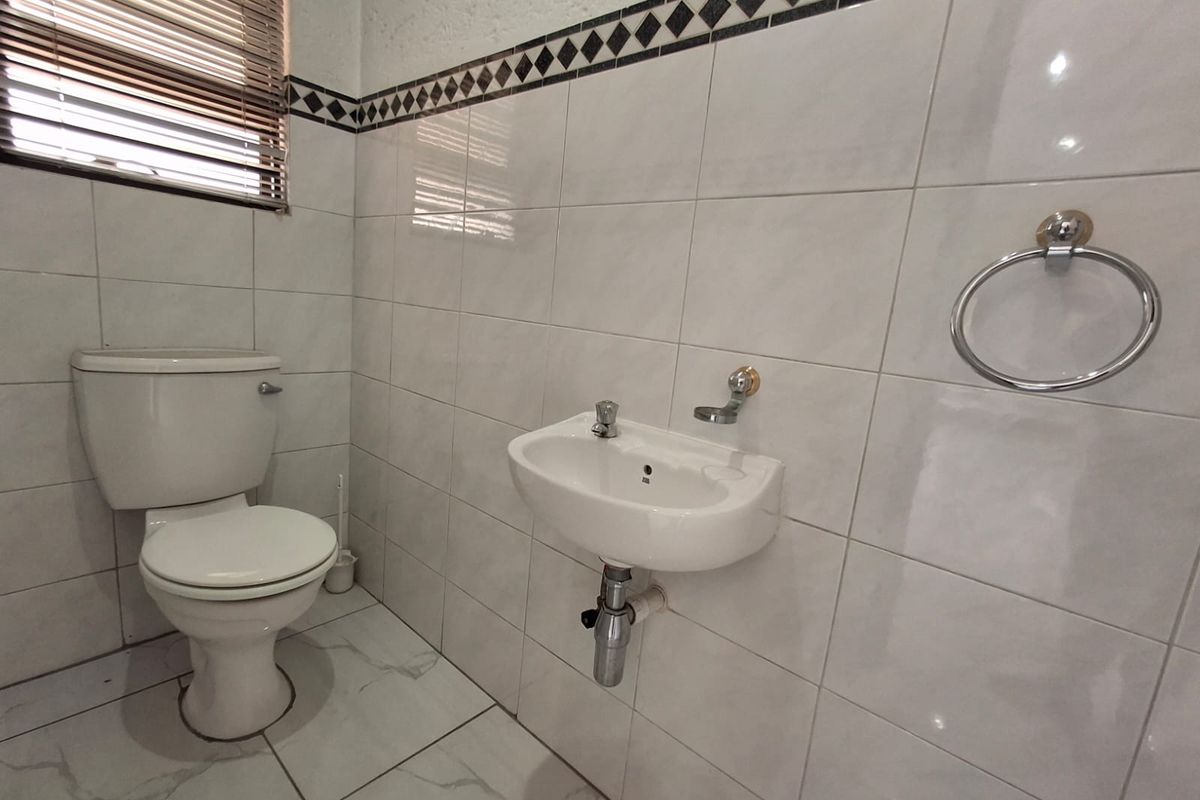 A functional guest bathroom featuring white wall and floor tiles, a wall-mounted basin, and a standard toilet with a window blind.