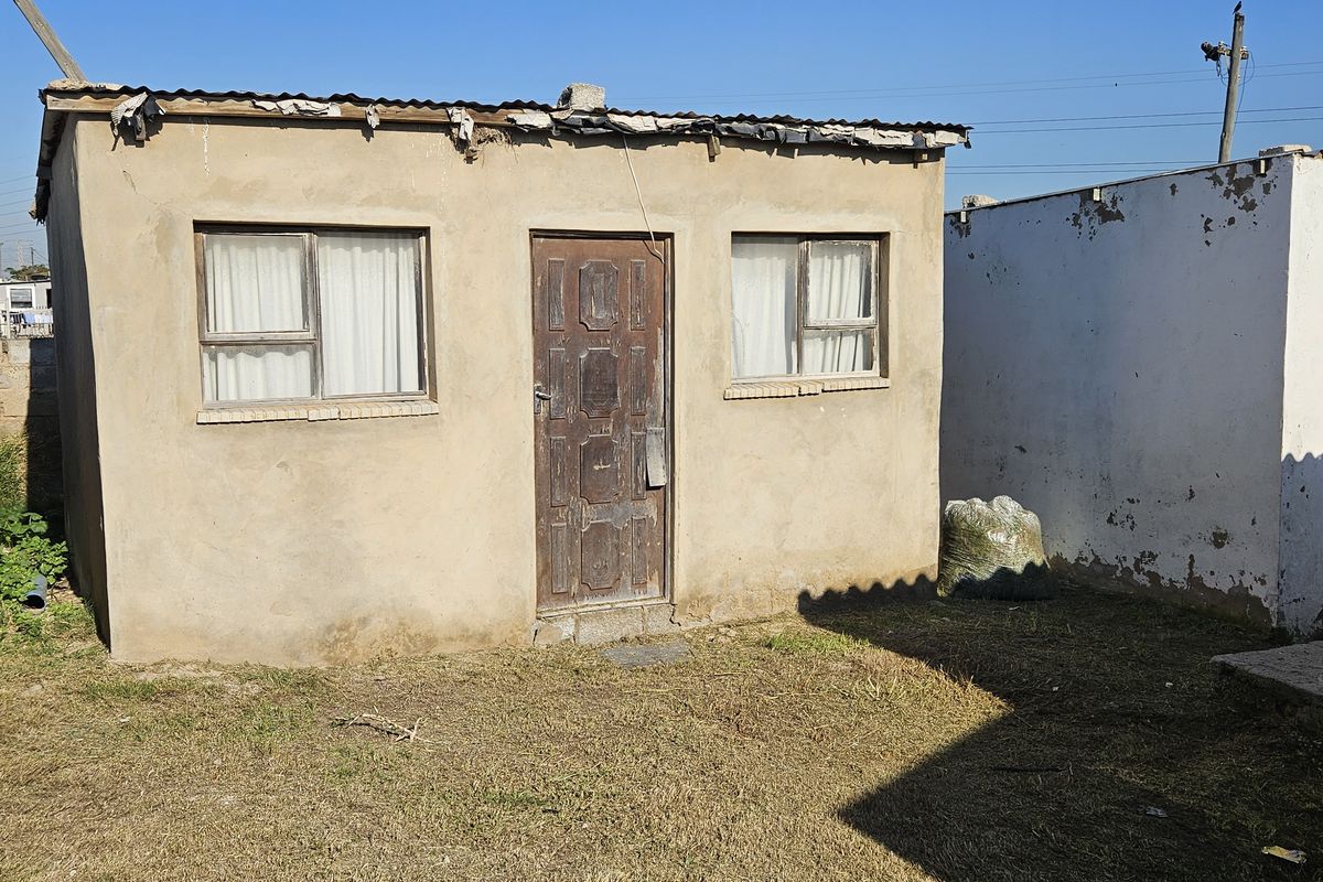 Rear exterior view of a small townhouse unit featuring a corrugated metal roof and basic plaster finish, showing signs of significant weathering.
