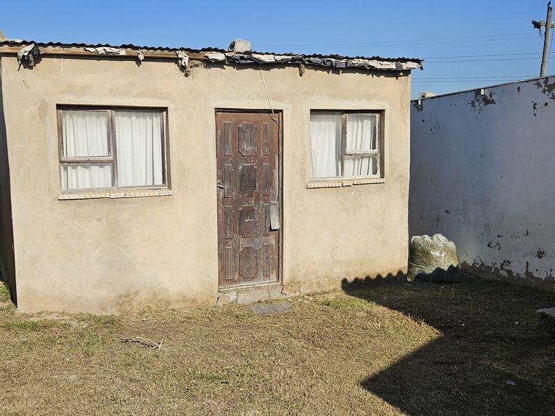 Rear exterior view of a small townhouse unit featuring a corrugated metal roof and basic plaster finish, showing signs of significant weathering.