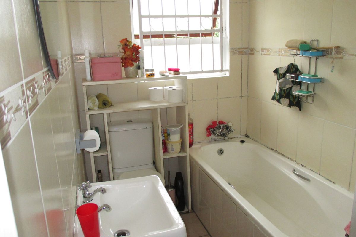 A compact bathroom featuring a built-in bathtub, pedestal washbasin, and toilet, finished with tiled walls and a window with security bars.