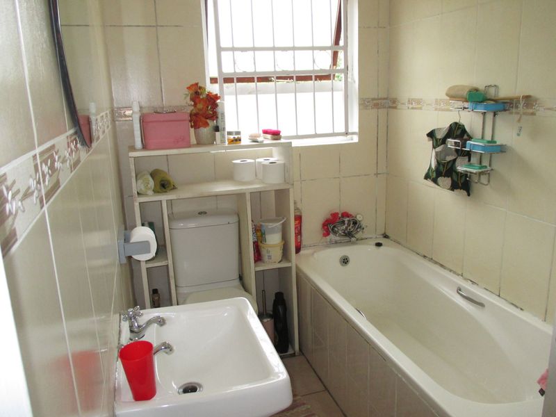 A compact bathroom featuring a built-in bathtub, pedestal washbasin, and toilet, finished with tiled walls and a window with security bars.