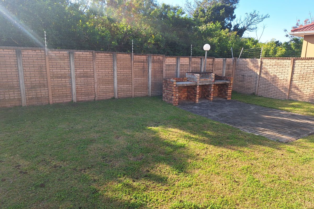 A private rear garden featuring a lawn, paved patio area, and a built-in brick braai, enclosed by a brick boundary wall with electric fencing.