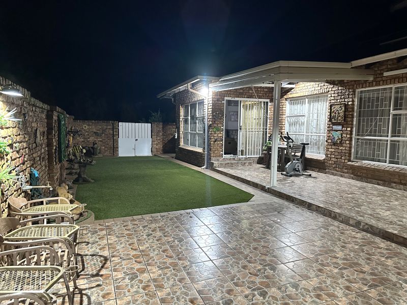 A night view of a rear exterior area featuring a covered tiled patio, brick wall construction, and an artificial lawn garden space.