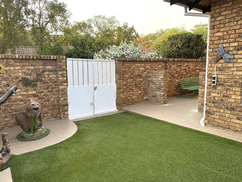 A private rear courtyard featuring a face brick boundary wall, tiled patio area, and low-maintenance artificial lawn with a secure double gate.