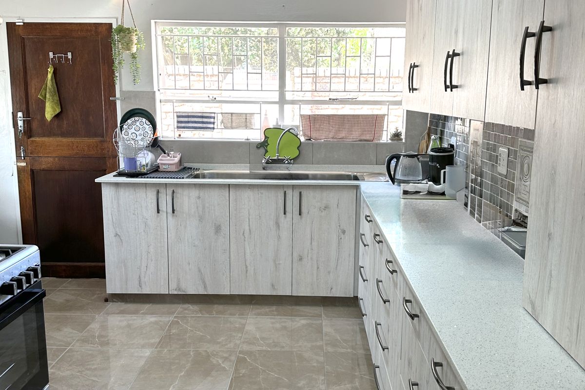 A functional kitchen featuring light wood-grain cabinetry, stone countertops, tiled flooring, and a large window with security bars, providing ample storage and workspace.