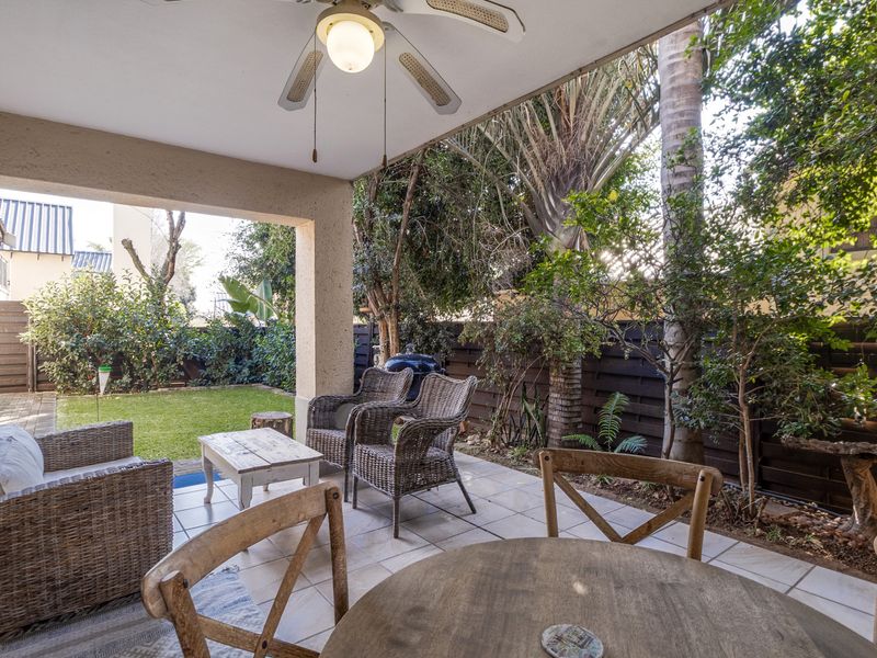 A covered outdoor patio area featuring tiled flooring and a ceiling fan, overlooking a private garden with established trees and timber boundary fencing.