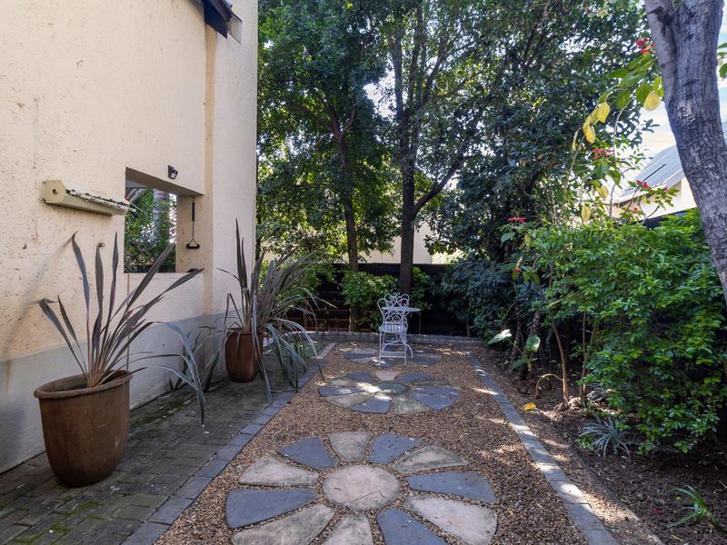 A private rear garden featuring a decorative stone path, mature trees, and established landscaping alongside the townhouse exterior wall.