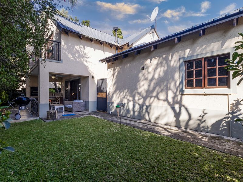 Rear view of the townhouse featuring a private garden, paved walkway, covered patio with built-in braai, and an upper-level balcony.