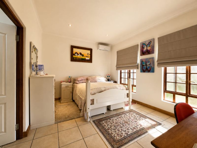 A well-lit bedroom featuring tiled flooring, a wall-mounted air conditioning unit, and wooden-framed windows with fitted roman blinds.