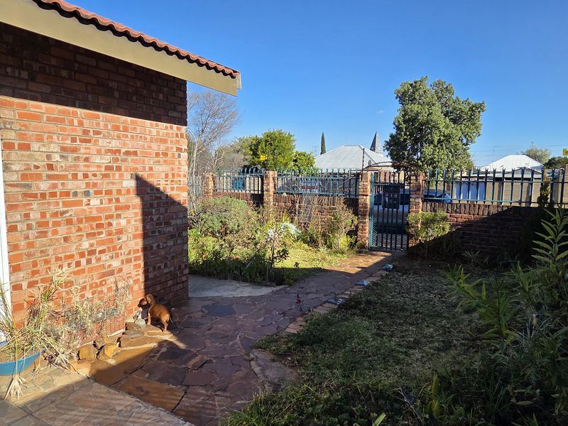 A rear exterior view showing a brick house facade, paved walkway, and enclosed garden area with perimeter wall and metal fencing.