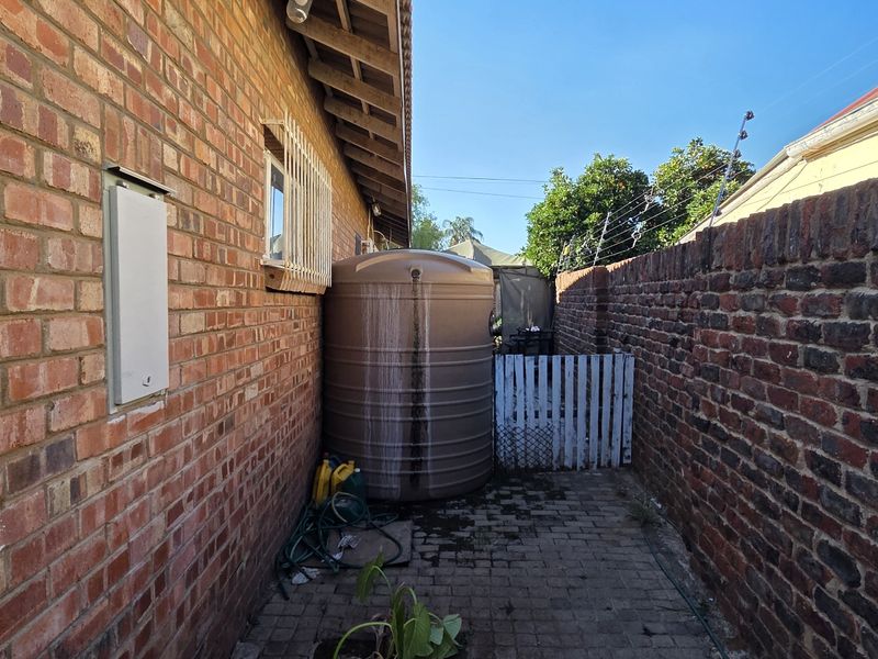 A narrow side passage featuring a brick boundary wall, paved flooring, and a large water storage tank with security fencing.