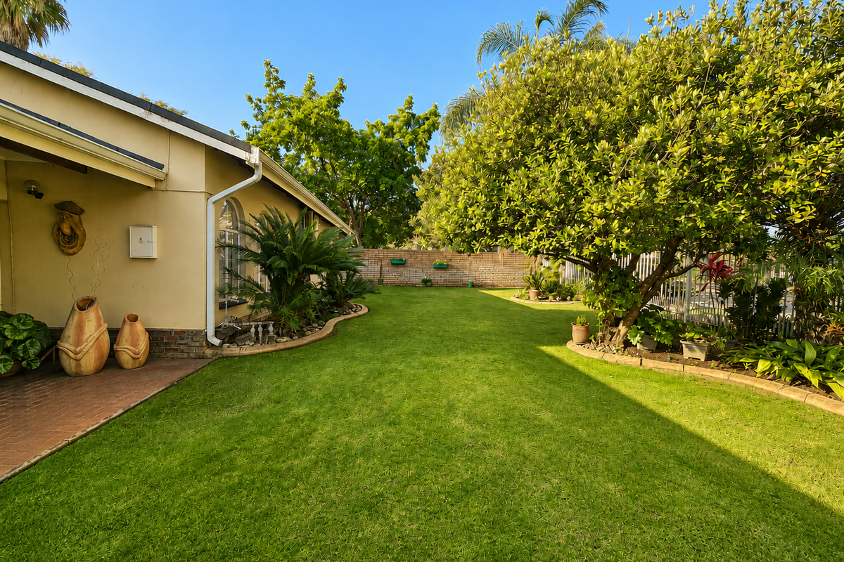 A well-maintained rear garden featuring a paved patio area, lush lawn, mature trees, and established garden beds enclosed by a brick boundary wall.