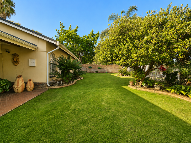 A well-maintained rear garden featuring a paved patio area, lush lawn, mature trees, and established garden beds enclosed by a brick boundary wall.