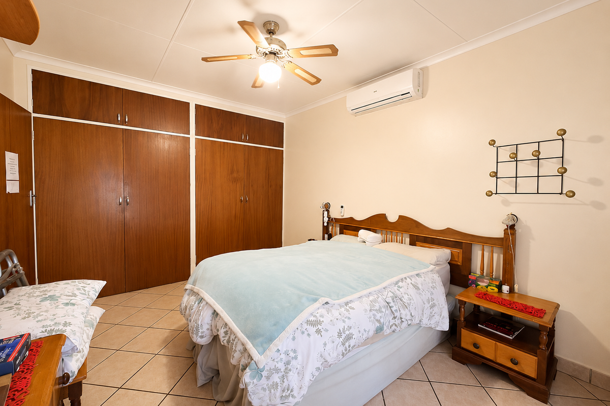 A bedroom featuring built-in wooden cupboards, a ceiling fan, and a wall-mounted split-system air conditioner with tiled flooring throughout.