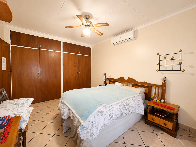 A bedroom featuring built-in wooden cupboards, a ceiling fan, and a wall-mounted split-system air conditioner with tiled flooring throughout.