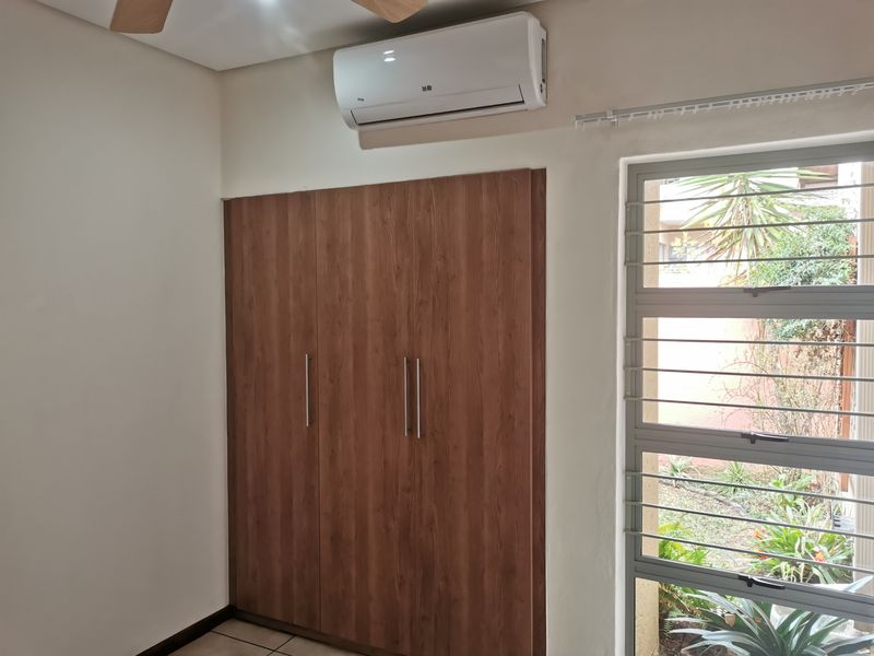 A clean bedroom featuring a built-in wood-finish wardrobe, a wall-mounted split-system air conditioner, and a large window with security bars overlooking the garden.
