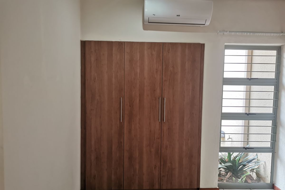 A clean bedroom featuring a built-in wooden wardrobe, a wall-mounted air conditioning unit, and a window with security bars overlooking a small garden area.