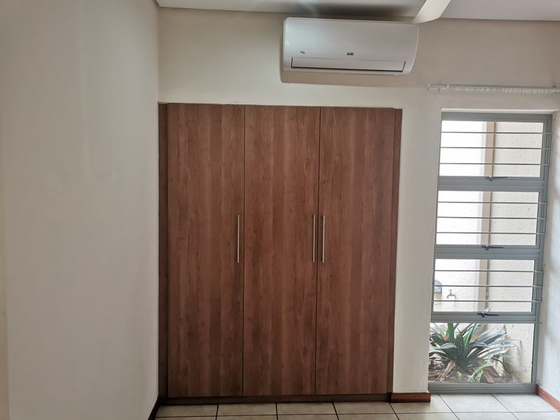 A clean bedroom featuring a built-in wooden wardrobe, a wall-mounted air conditioning unit, and a window with security bars overlooking a small garden area.