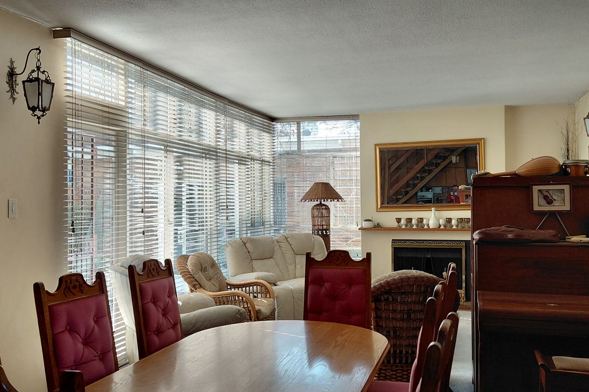 A spacious living area featuring a fireplace with a mantelpiece, large windows with venetian blinds, and classic wall-mounted lighting fixtures.