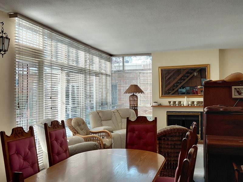 A spacious living area featuring a fireplace with a mantelpiece, large windows with venetian blinds, and classic wall-mounted lighting fixtures.