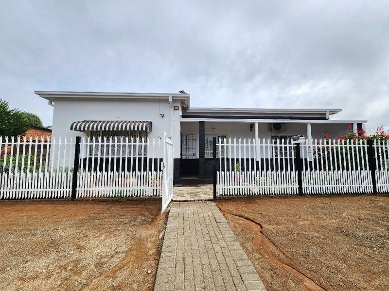 Front elevation of a single-story house featuring a white picket fence, paved entrance walkway, and security features including window bars and an awning.