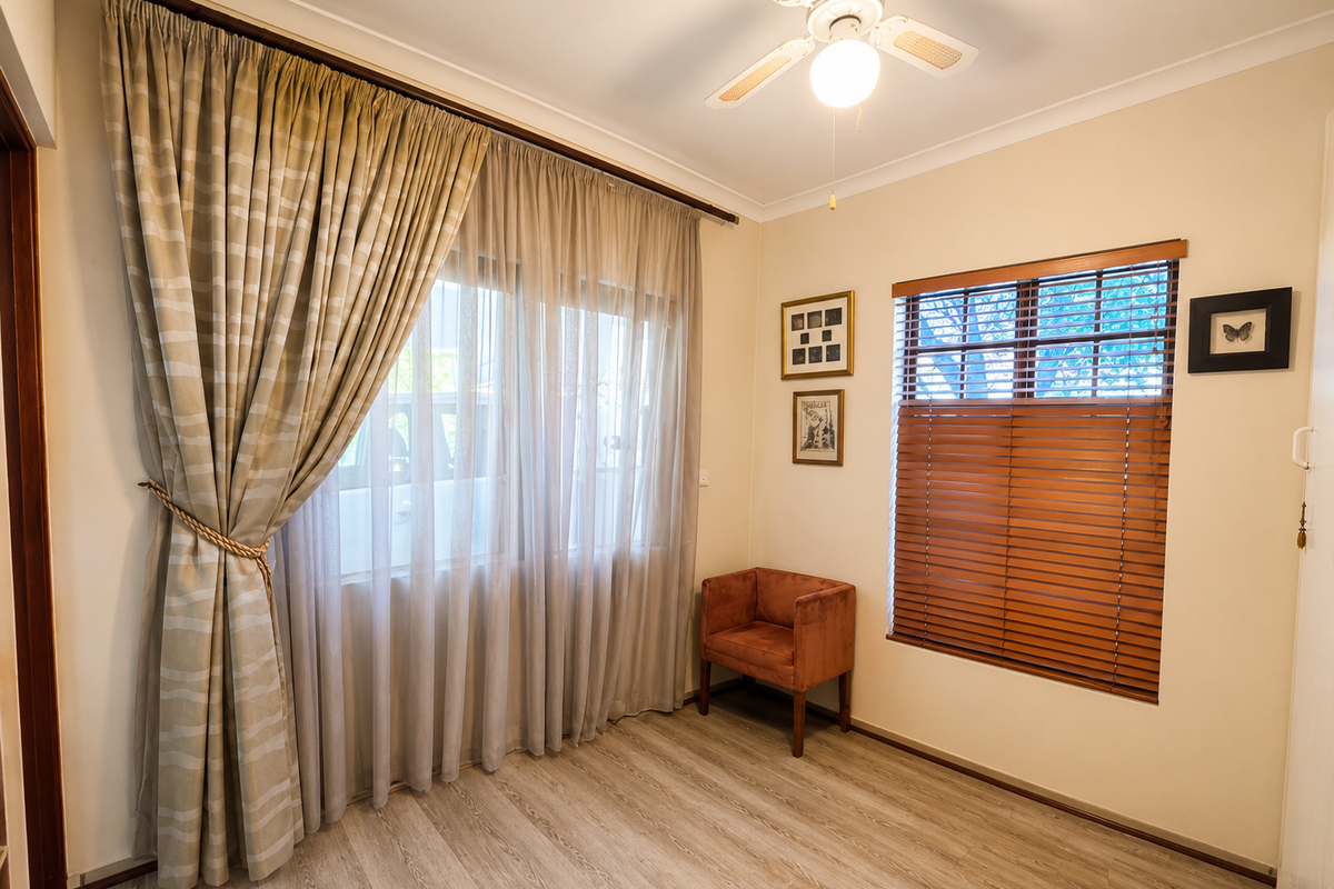 A well-maintained bedroom featuring light-toned laminate flooring, a ceiling fan, and a combination of sheer curtains and wooden venetian blinds.