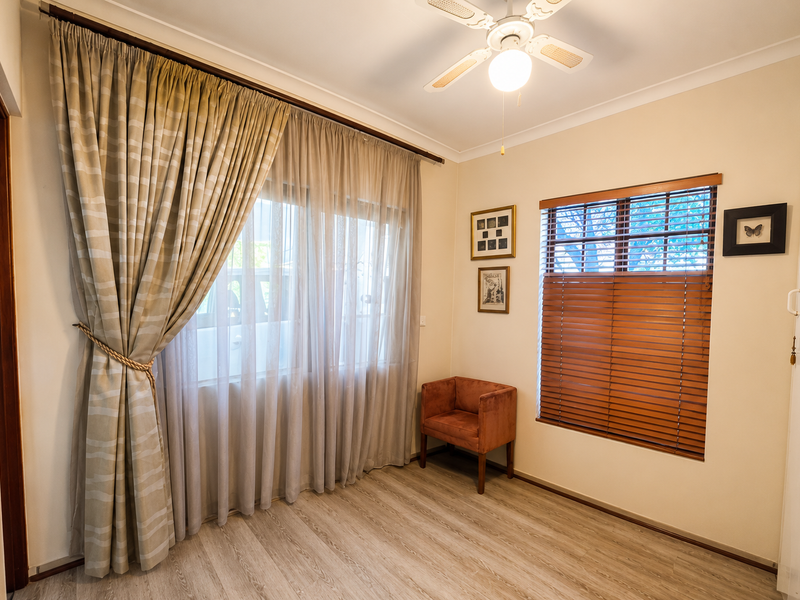 A well-maintained bedroom featuring light-toned laminate flooring, a ceiling fan, and a combination of sheer curtains and wooden venetian blinds.