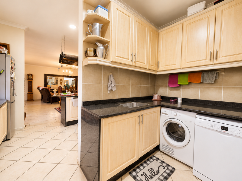 A functional scullery area featuring light wood cabinetry, dark granite countertops, a stainless steel sink, and integrated laundry appliances, adjacent to the main living space.