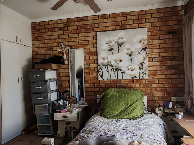 A bedroom featuring an exposed brick feature wall, built-in cupboards, and a ceiling fan, offering a functional space with potential for cosmetic updates.