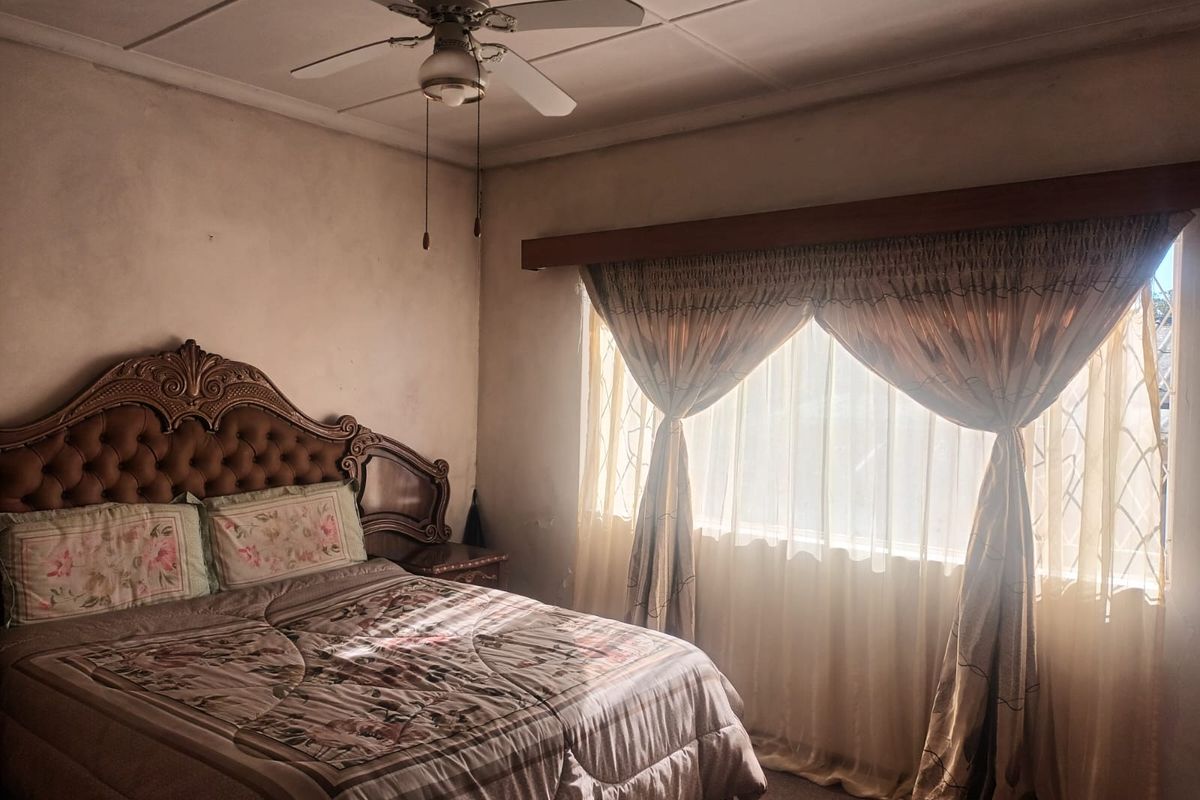 A bedroom featuring a ceiling fan and large window with curtain rod, showing signs of wear on the wall surfaces.