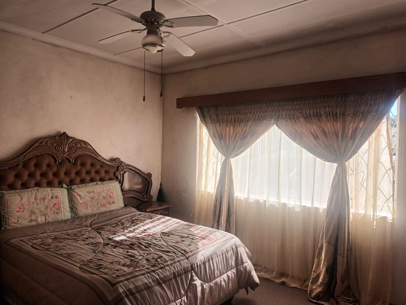 A bedroom featuring a ceiling fan and large window with curtain rod, showing signs of wear on the wall surfaces.