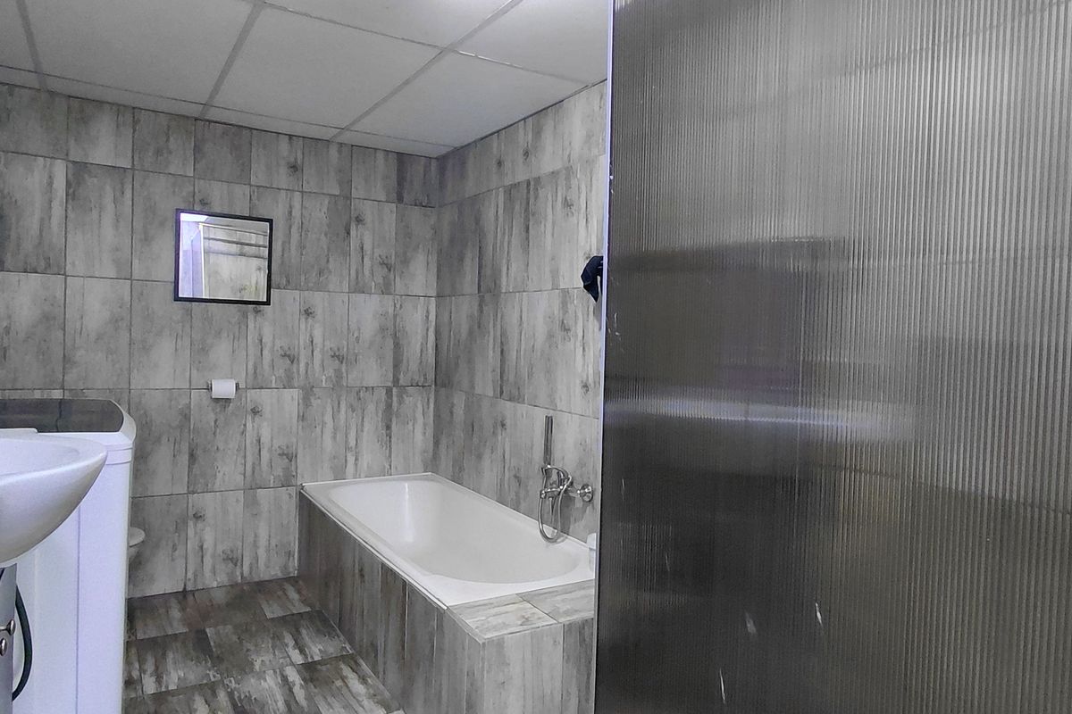 A functional bathroom featuring grey wood-look wall and floor tiles, a built-in bathtub, and a wall-mounted basin with integrated laundry space.