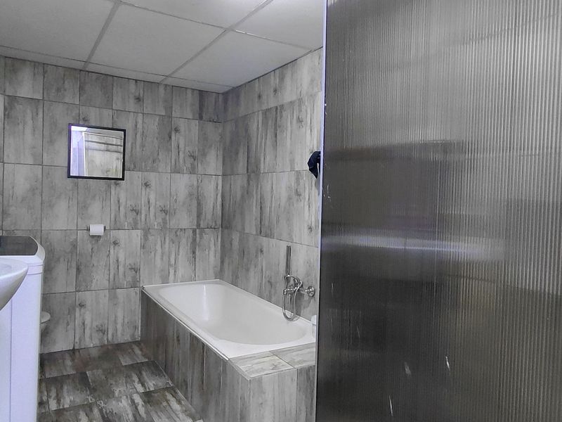 A functional bathroom featuring grey wood-look wall and floor tiles, a built-in bathtub, and a wall-mounted basin with integrated laundry space.