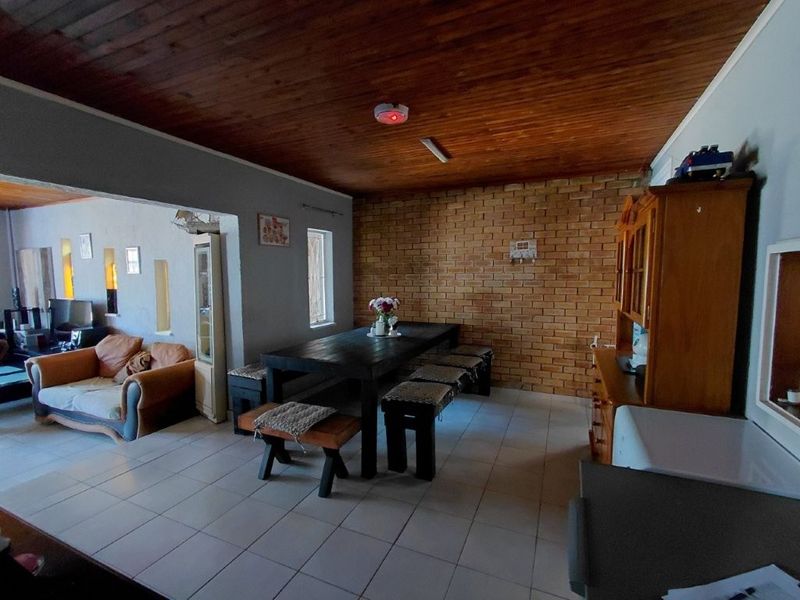 Open plan living and dining area featuring a rustic exposed brick wall, timber ceiling, and neutral ceramic floor tiles.