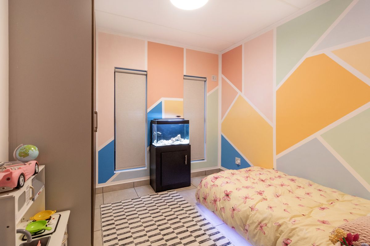 A bright bedroom featuring tiled flooring, built-in wardrobe storage, and dual windows with roller blinds, finished with a vibrant geometric accent wall.