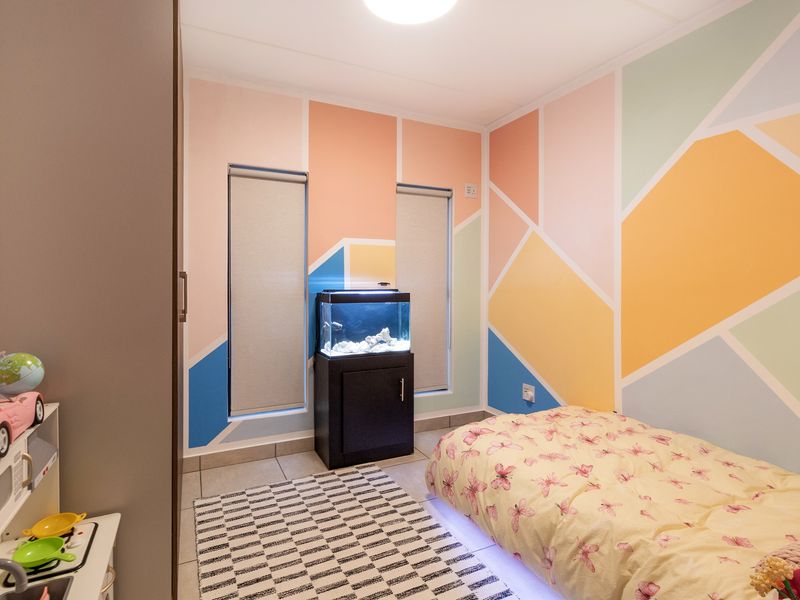 A bright bedroom featuring tiled flooring, built-in wardrobe storage, and dual windows with roller blinds, finished with a vibrant geometric accent wall.