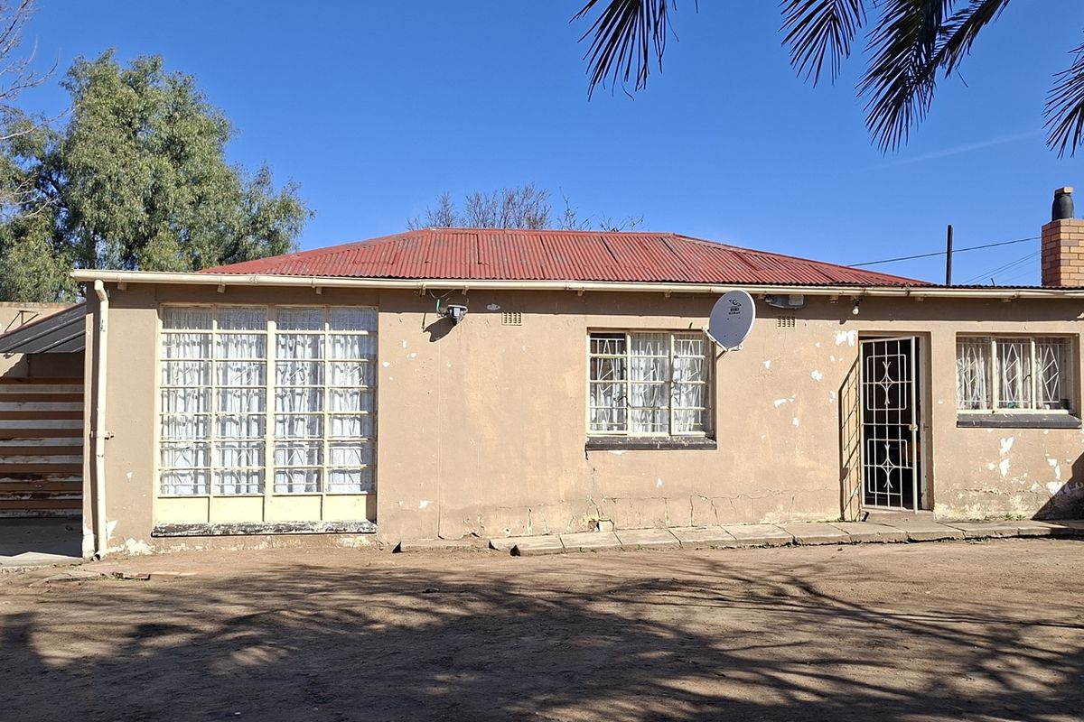 Rear view of a single-story house featuring a corrugated metal roof, brick chimney, and security-fitted windows, set against a large open yard area.