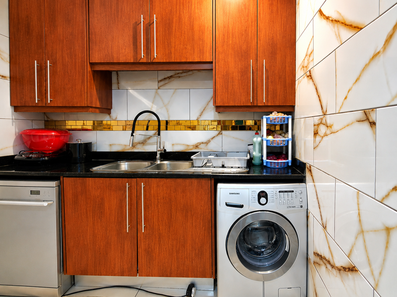 A functional scullery featuring wood-finish cabinetry, black granite countertops, a stainless steel double sink, and integrated laundry and dishwashing appliances.