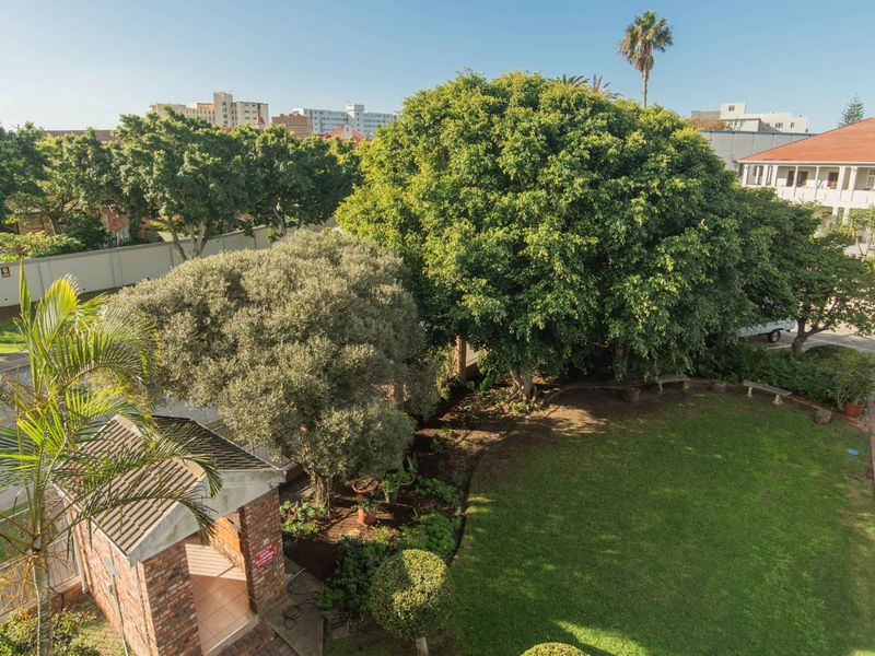 An elevated view overlooking a well-maintained communal garden area with mature trees, brick structures, and paved pathways within the apartment complex grounds.