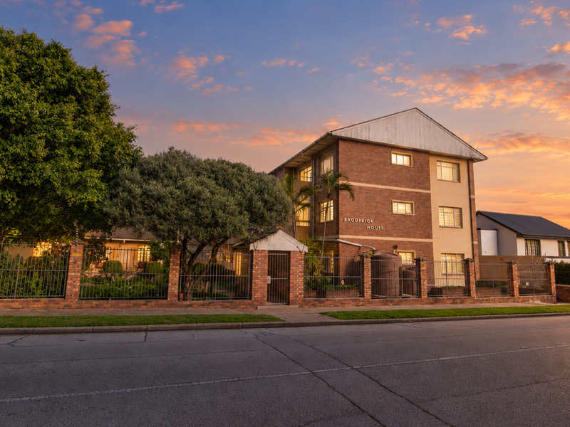 Street-facing view of Broderick House, a multi-story brick apartment building featuring secure perimeter fencing and established landscaping at sunset.