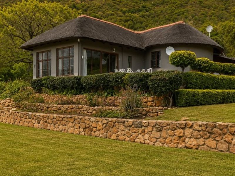 A charming farmhouse featuring a traditional thatched roof, set against a lush hillside with tiered stone retaining walls and a well-maintained garden.