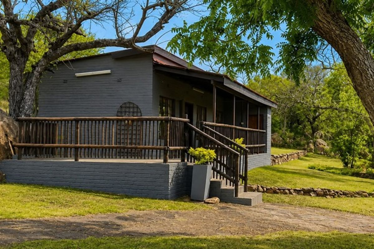 A charming farm cottage featuring a painted brick exterior, a raised timber deck with railing, and a scenic, natural garden setting.