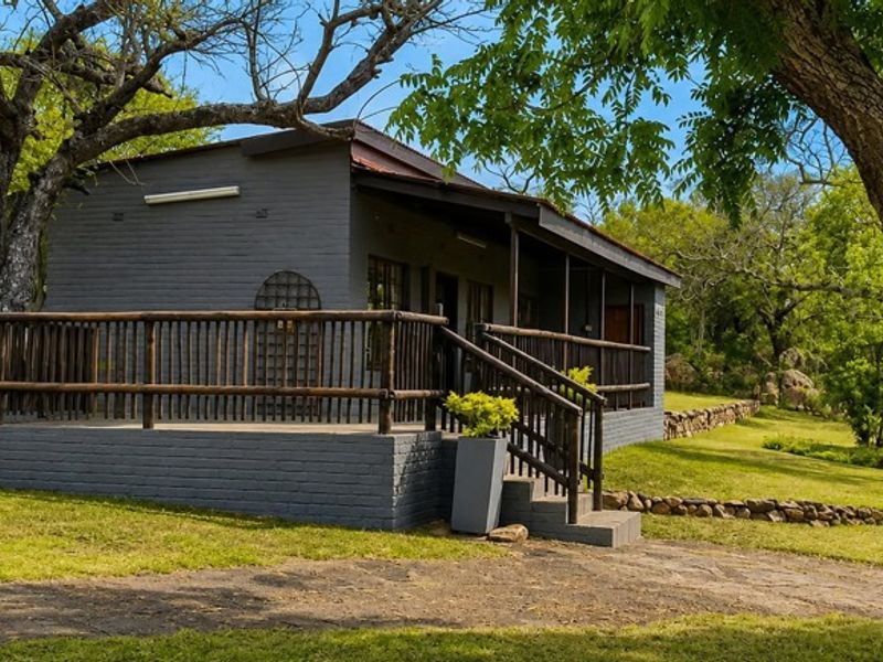 A charming farm cottage featuring a painted brick exterior, a raised timber deck with railing, and a scenic, natural garden setting.