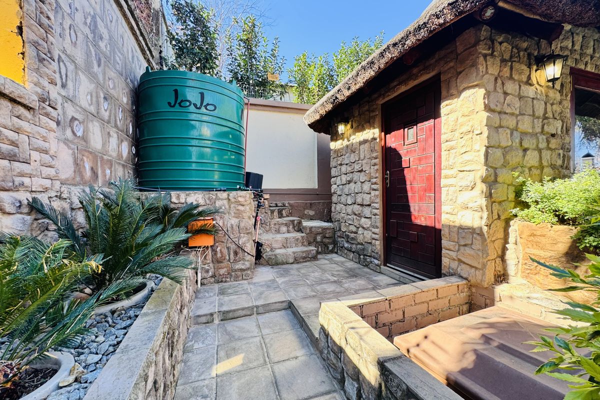 A well-maintained rear exterior area featuring stone-clad walls, a thatched roof structure, a paved pathway, and a large water storage tank.
